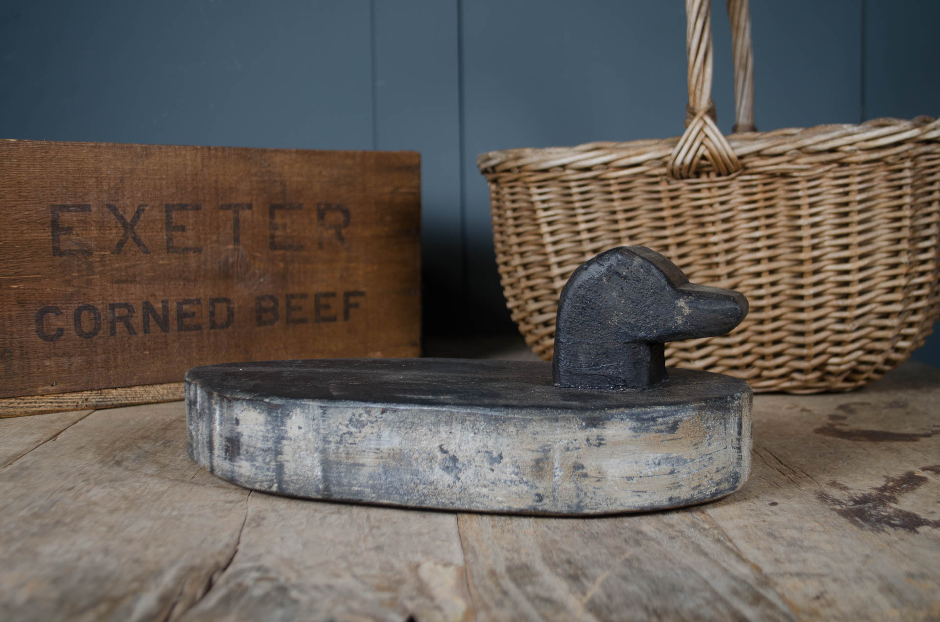
A rustic wooden table display featuring a dark metal and weathered wood object in the foreground, with a woven wicker basket and a wooden sign reading 'EXETER CORNED BEEF' in the background against a dark blue paneled wall.