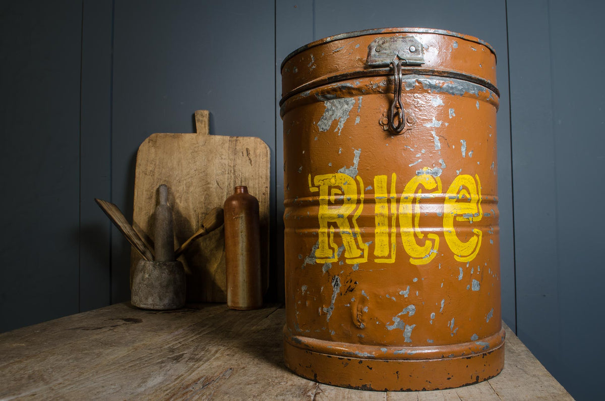 A vintage rusted orange metal rice canister with yellow 'RICE' lettering, displayed on a rustic wooden table next to a wooden cutting board and small metal containers against a dark blue paneled wall.