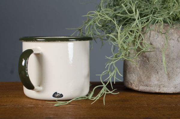 White mug with black handle on a wooden surface next to a potted plant against a gray wall.