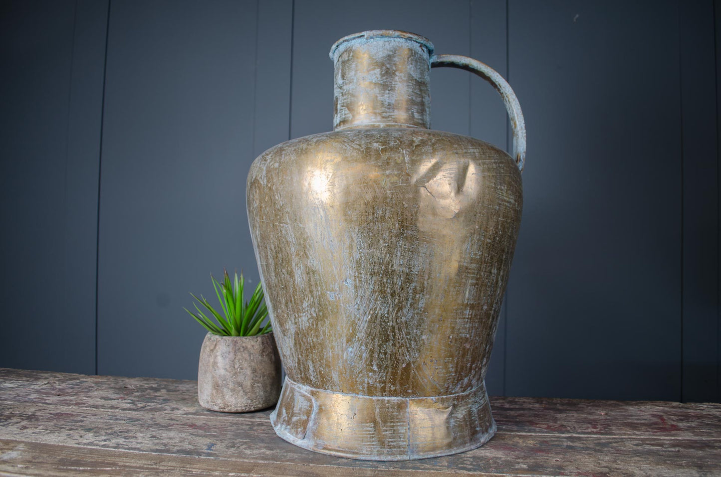 Large antique brass water jug with weathered verdigris patina, polished finish, and handle, displayed on rustic wooden surface.