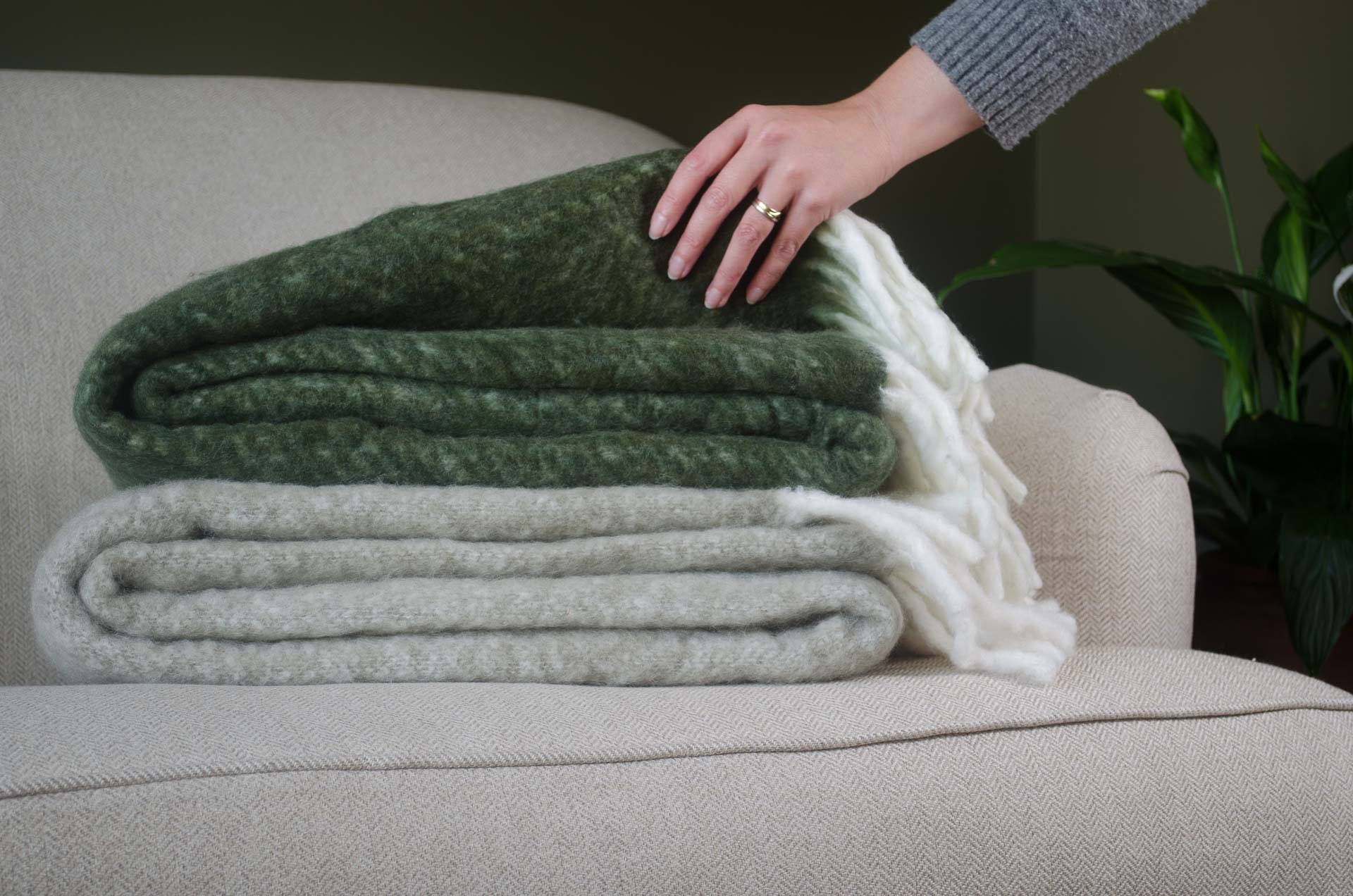 Person arranging folded lambs wool tail blankets on a couch with a plant in the background