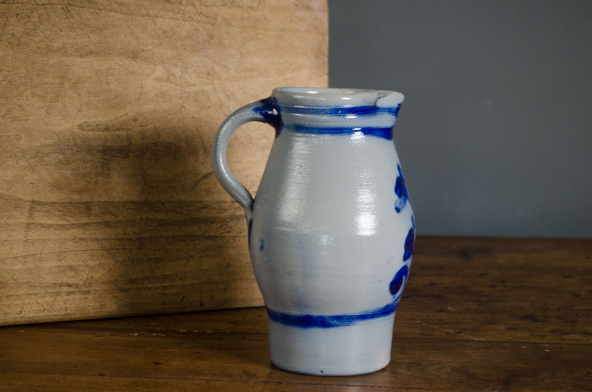 
A handmade ceramic pitcher with a white glaze and blue hand-painted floral patterns, set on a wooden table next to a wooden board.