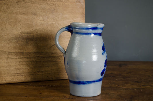 A handmade ceramic pitcher with a white glaze and blue hand-painted floral patterns, set on a wooden table next to a wooden board.