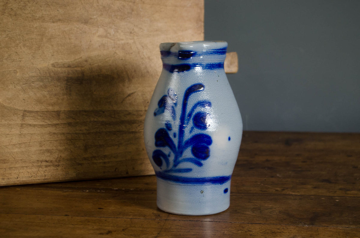 A small handmade ceramic bottle with a blue floral pattern on a white glazed body, standing on a wooden surface against a rustic wood and gray background.