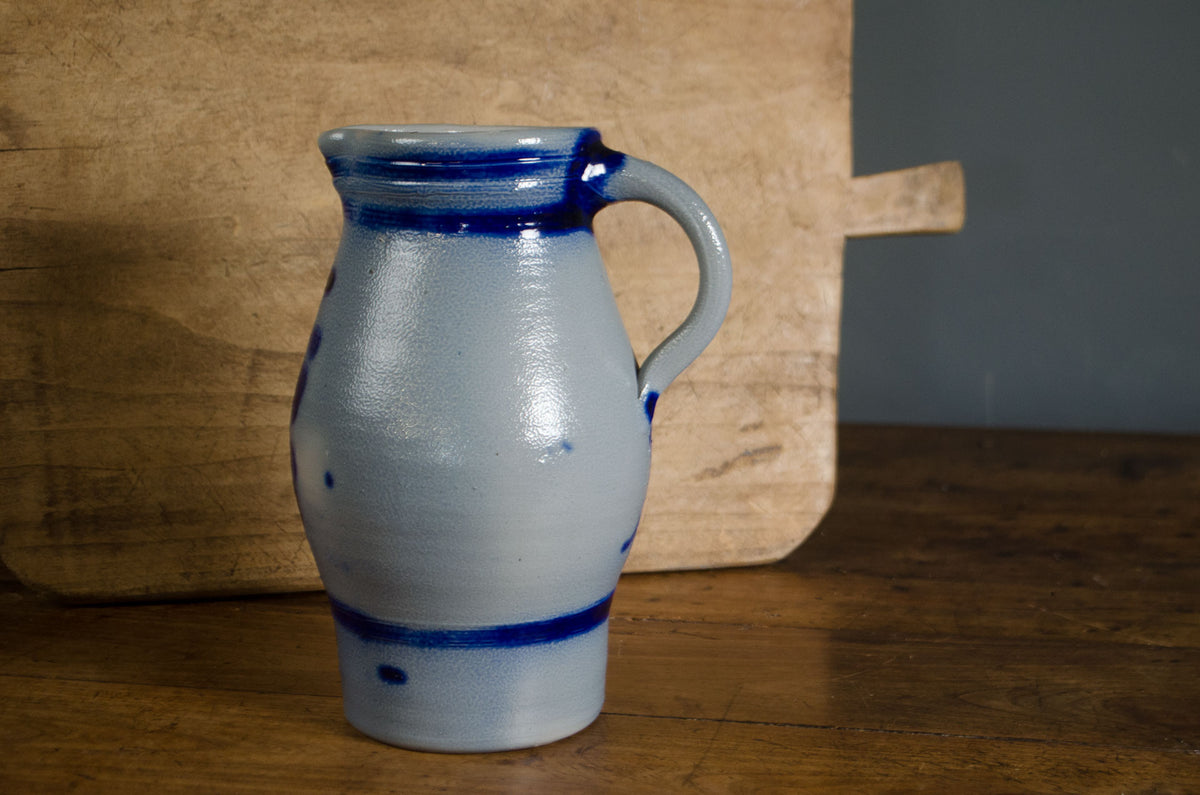 A small ceramic pitcher with a pale blue-gray glaze and dark cobalt blue bands and handle, standing on a wooden table with a rustic wooden board behind it.