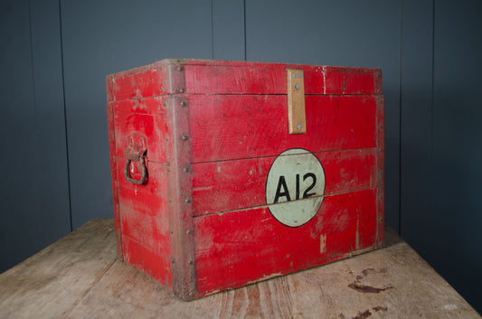 A vintage red wooden storage chest with metal rivets, a leather handle, and a circular white label reading 'A12', displayed on a rustic wooden table against a dark paneled wall.