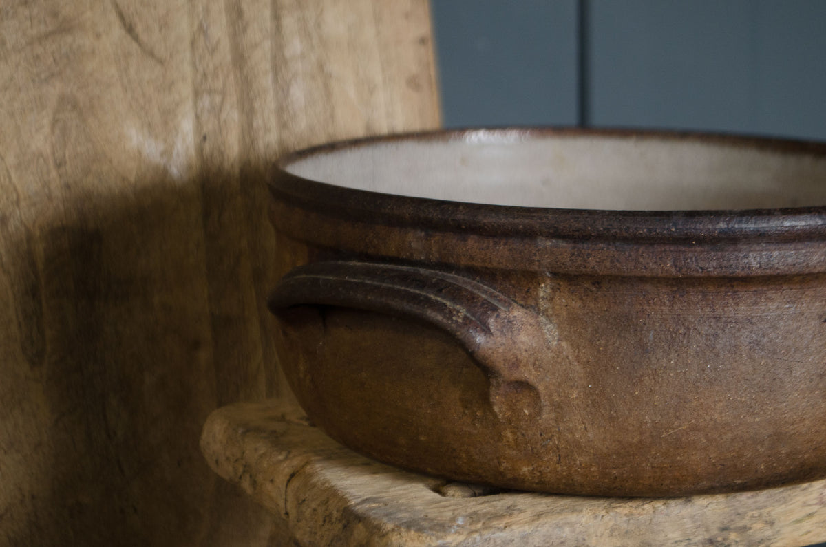 A rustic brown ceramic pot with a dark rim and a light beige interior, shown close-up on a wooden surface against a wood and blue-gray background.