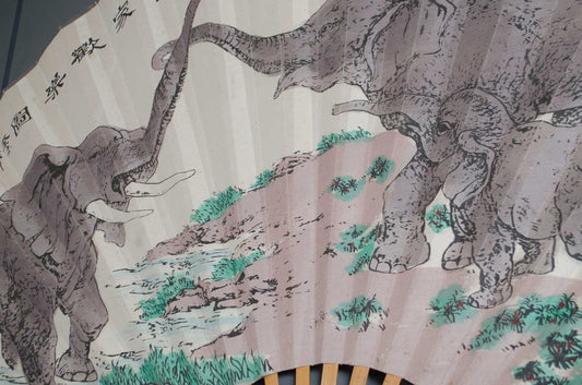 A folding paper fan featuring a traditional Chinese-style illustration of three elephants in gray and black ink with green foliage and a beige landscape background.