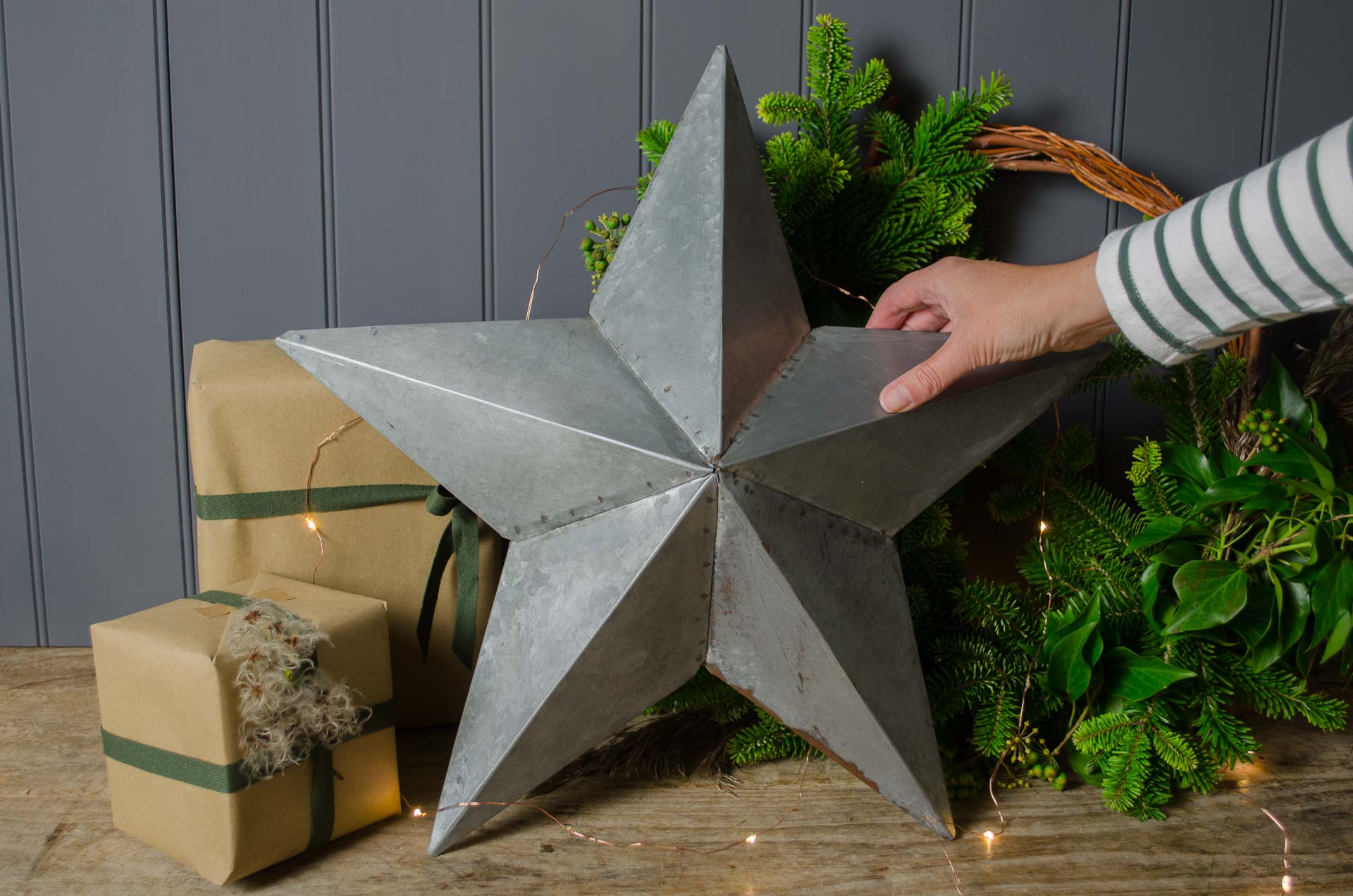 Hand placing a large reclaimed metal barn star among wrapped gifts and evergreen decorations on a wooden table