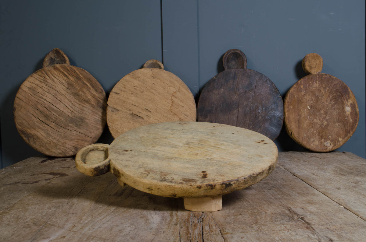 A rustic round wooden serving board with a small loop handle sits on a weathered wooden table, with four larger wooden cutting or serving boards leaning against a dark blue-gray paneled wall in the background.
