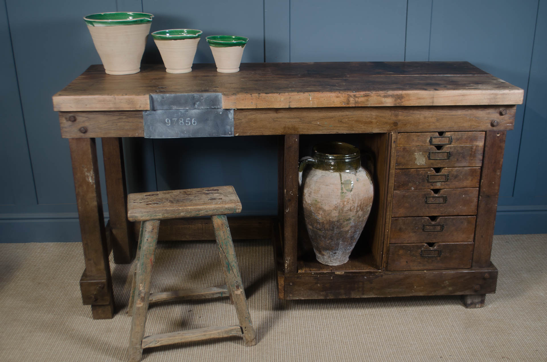 Vintage wooden desk with a stool and ceramic items against a blue wall.