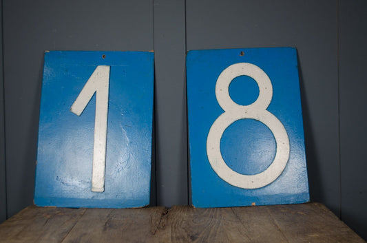 Two vintage blue wooden number signs displaying '1' and '8' side-by-side on a rustic wooden table against a dark gray paneled wall.
