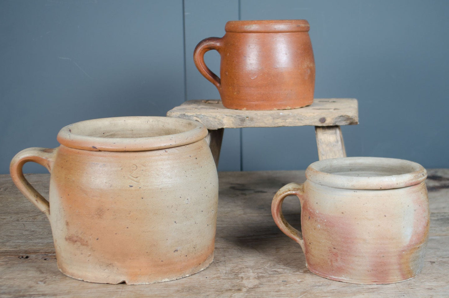 Three french stoneware rustic confit terracotta mugs on a wooden surface with a blue wall background