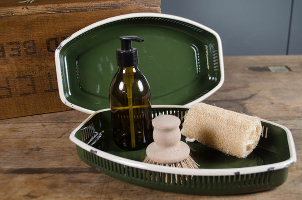 Green dish with cleaning supplies including a bottle, brush, and sponge on a wooden surface.