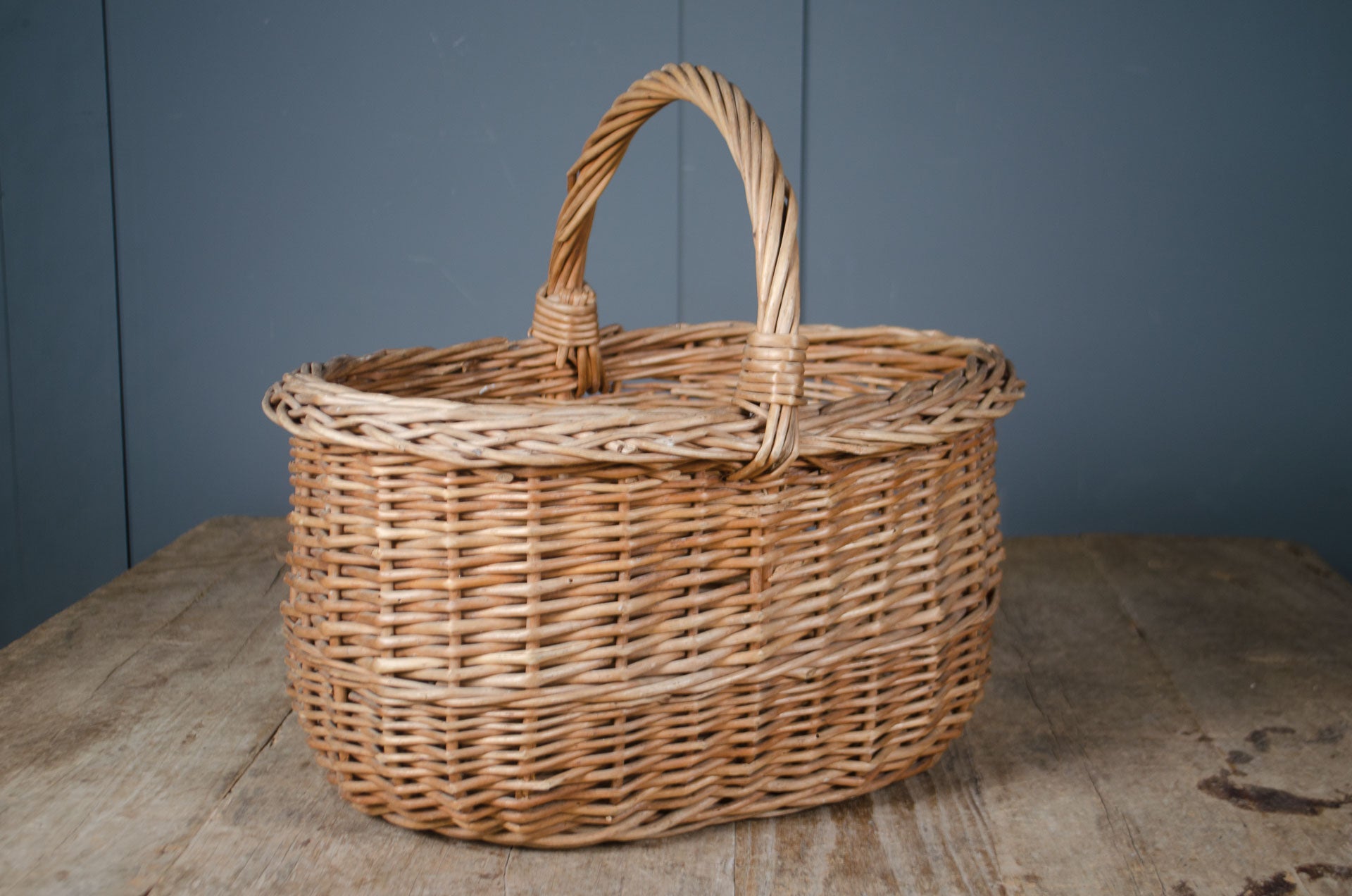 
A natural woven wicker basket with a single arched handle, placed on a rustic wooden surface against a dark blue-gray paneled background.