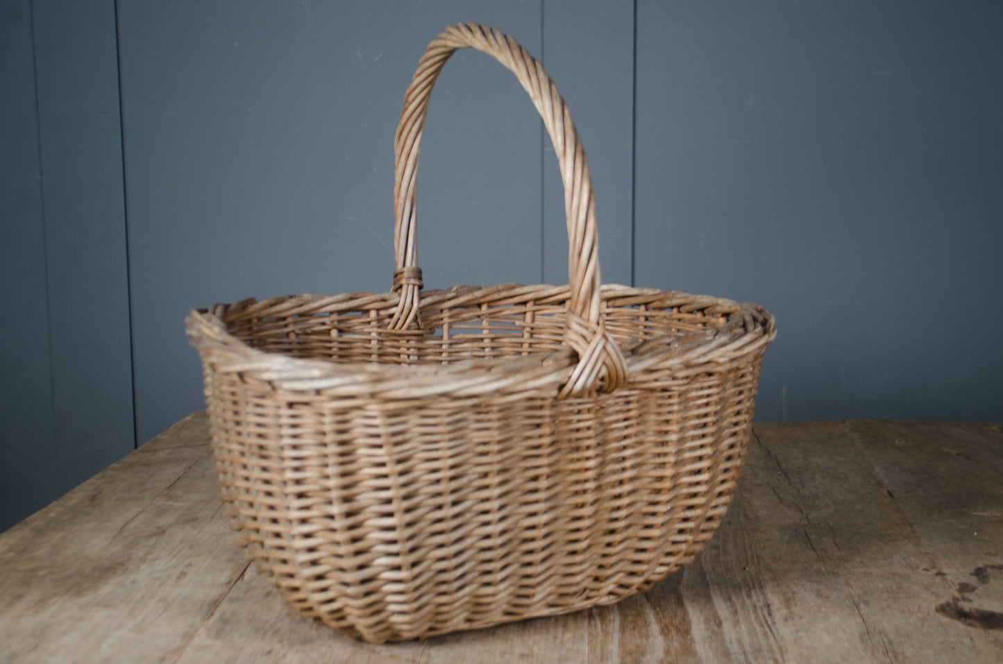 A natural woven wicker basket with a single twisted handle, set on a rustic wooden surface against a dark blue paneled wall.