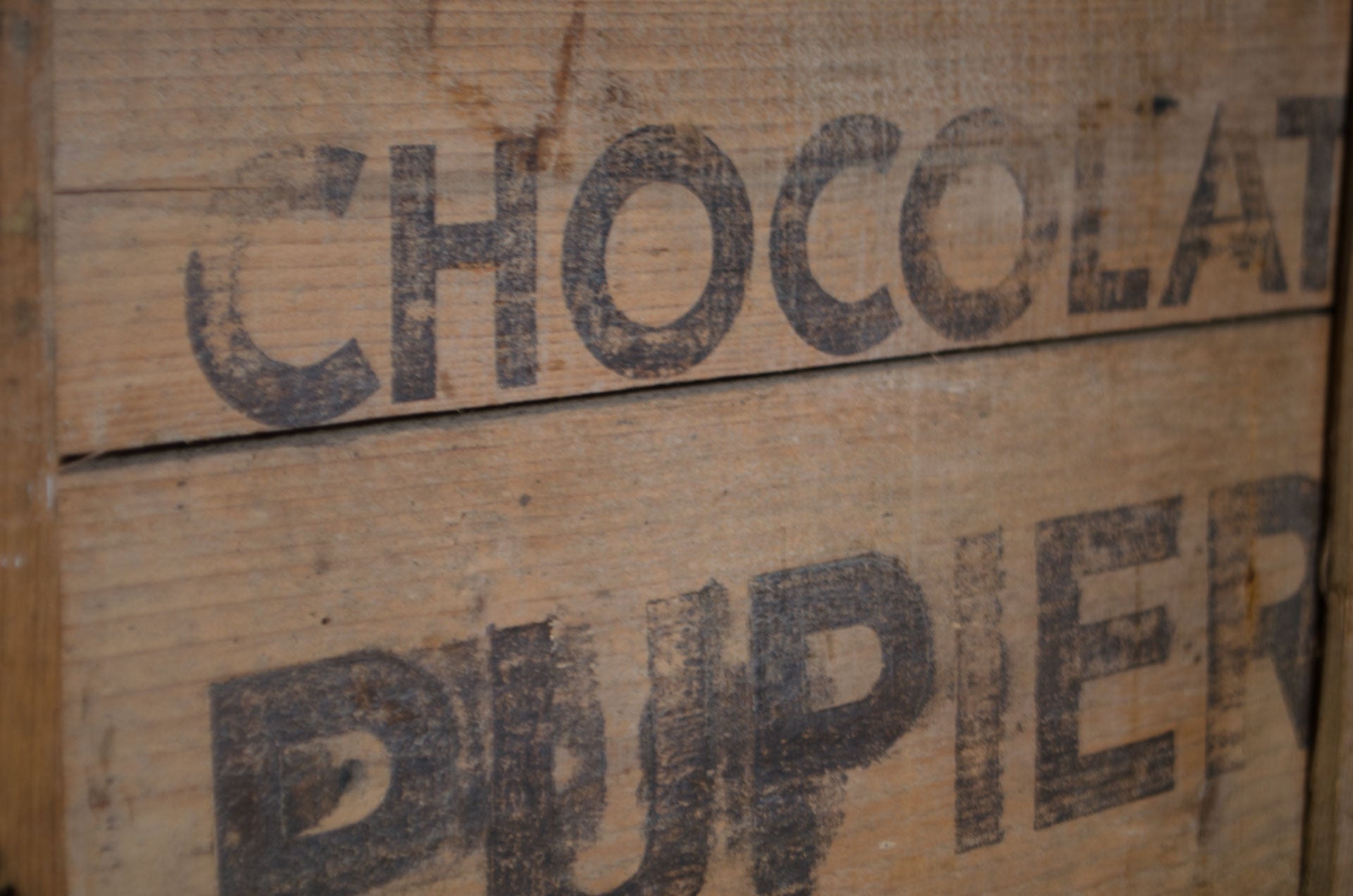 
Close-up of a rustic wooden box or crate with the words 'CHOCOLAT DU PIER' printed in dark stencil-style lettering on the front.