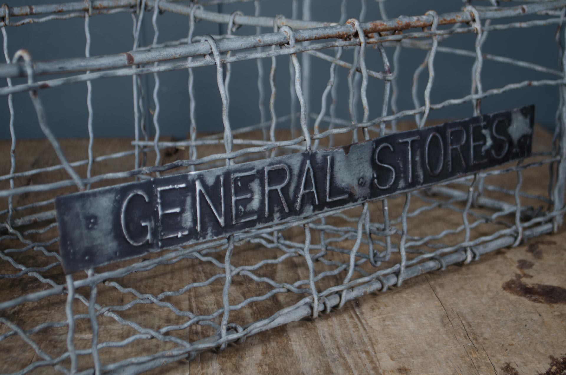 
A rustic metal sign reading 'GENERAL STORES' mounted on a galvanized wire cage, set against a dark blue-gray background and a wooden surface.