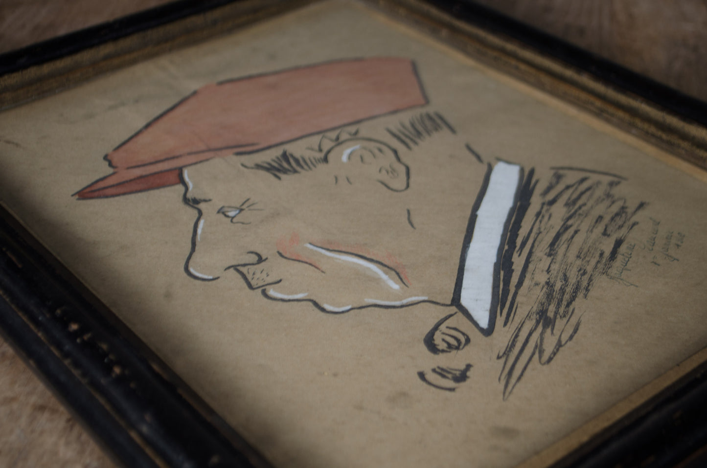 A framed portrait sketch of a man in profile wearing a reddish-brown hat, rendered in black and white line art on aged beige paper, shown in a dark wooden frame.