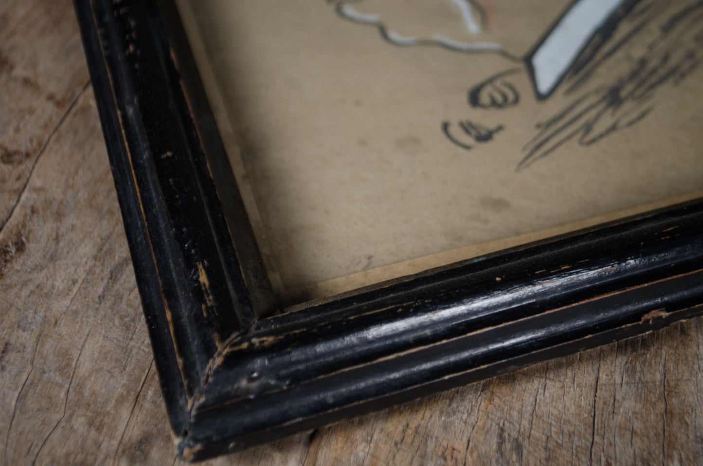 A close-up of a distressed black wooden picture frame corner with a beige-toned print or canvas inside, resting on a rustic wooden surface.