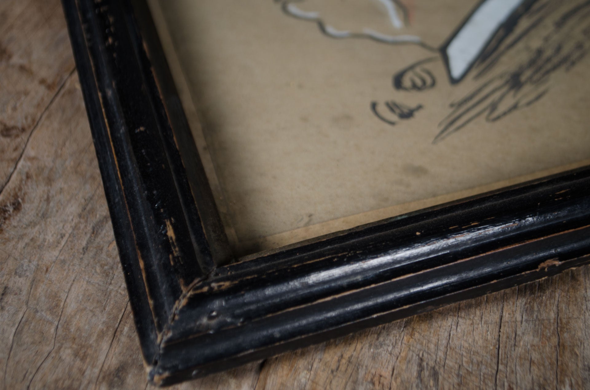 
A close-up of a distressed black wooden picture frame corner with a beige-toned print or canvas inside, resting on a rustic wooden surface.