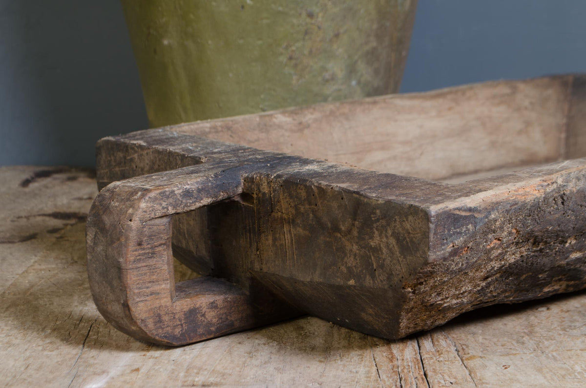 A rustic, hand-carved dark brown wooden kitchen bowl or trough with a looped handle, resting on a weathered wooden surface with a blurred olive-green ceramic pot in the background.