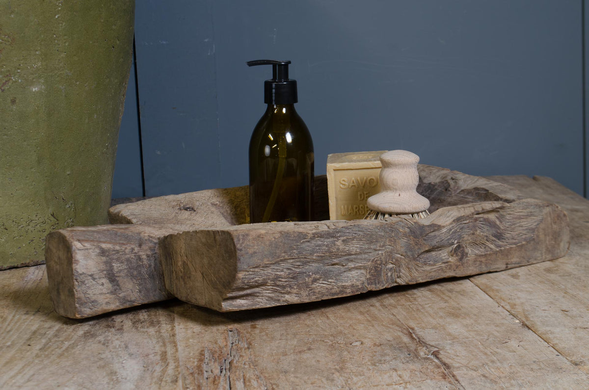 
A rustic carved wooden tray holding a dark amber pump bottle, a beige soap bar, and a small cream soap brush, set on a wooden surface against a blue-gray wall.
