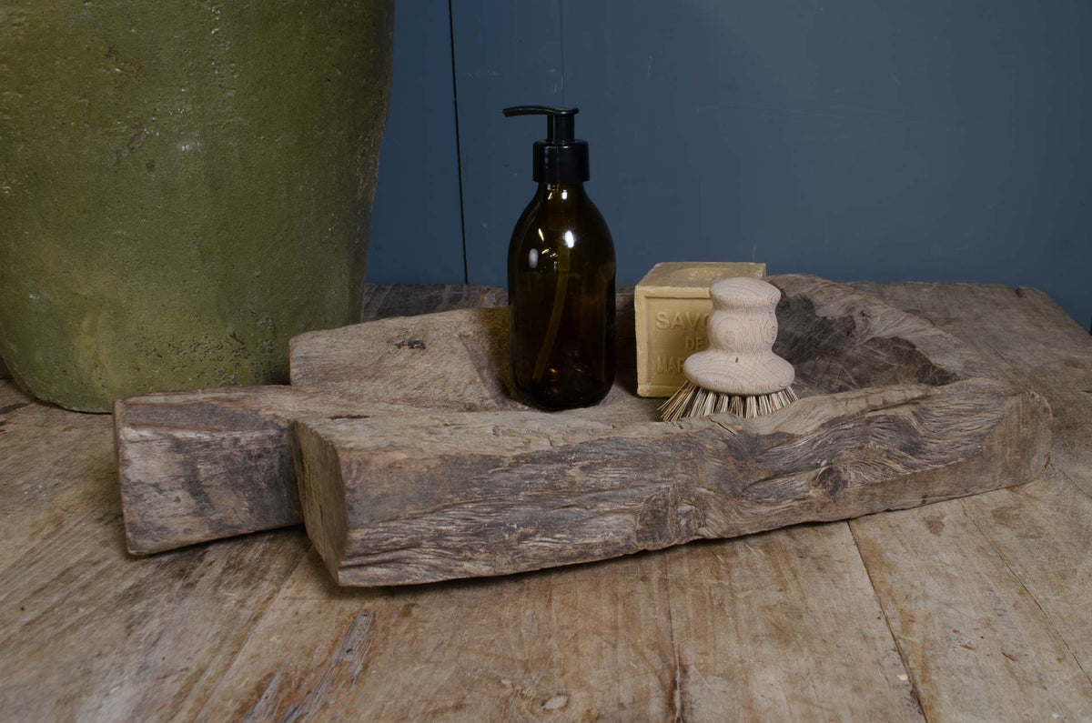 
A rustic carved wooden soap dish holding a dark amber pump bottle, a beige bar of soap, and a small wooden scrubber, set on a weathered wood surface against a blue-gray background.