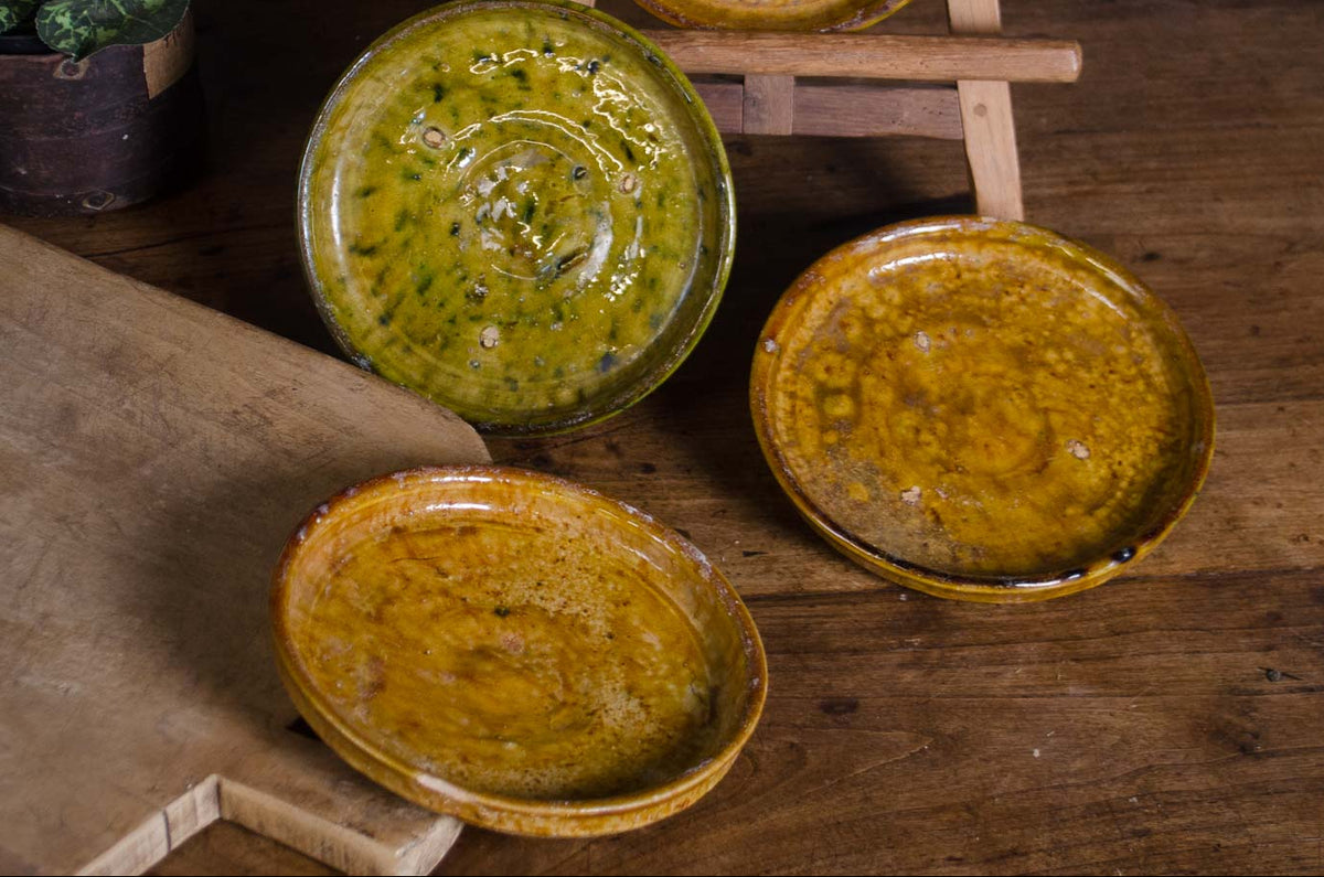 Four handmade ceramic plates with textured, rustic glazes in yellow, orange, and olive green, displayed on a wooden table with a wooden cutting board and a small potted plant.