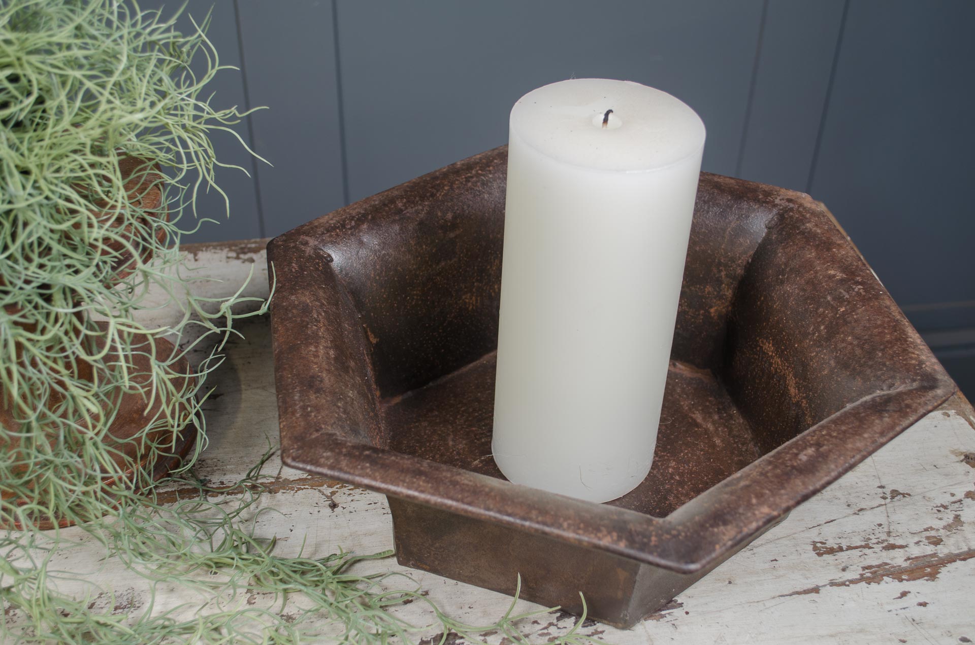 
White candle in a rustic metal holder on a wooden surface with plants in the background