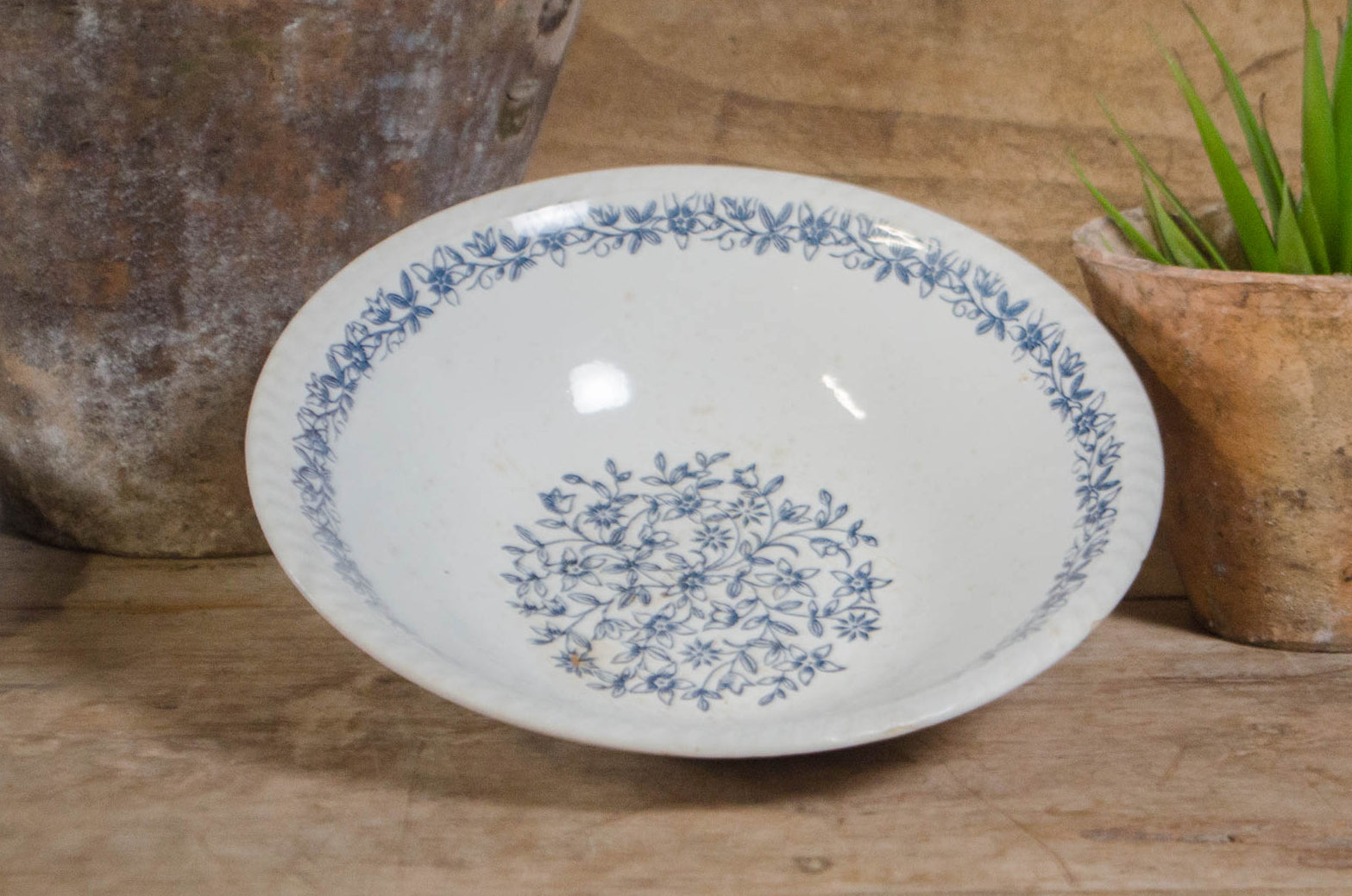 
A white ceramic bowl with a blue floral pattern, displayed on a rustic wooden surface next to a potted succulent and a large textured vase.