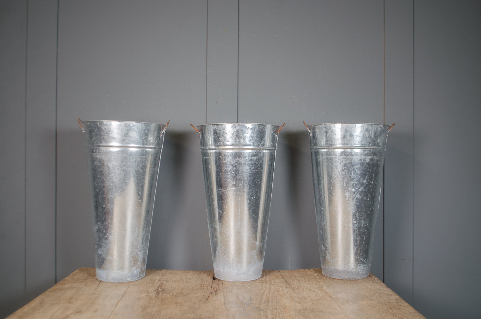 
Three galvanized metal buckets with side handles, standing on a wooden surface against a gray paneled wall.