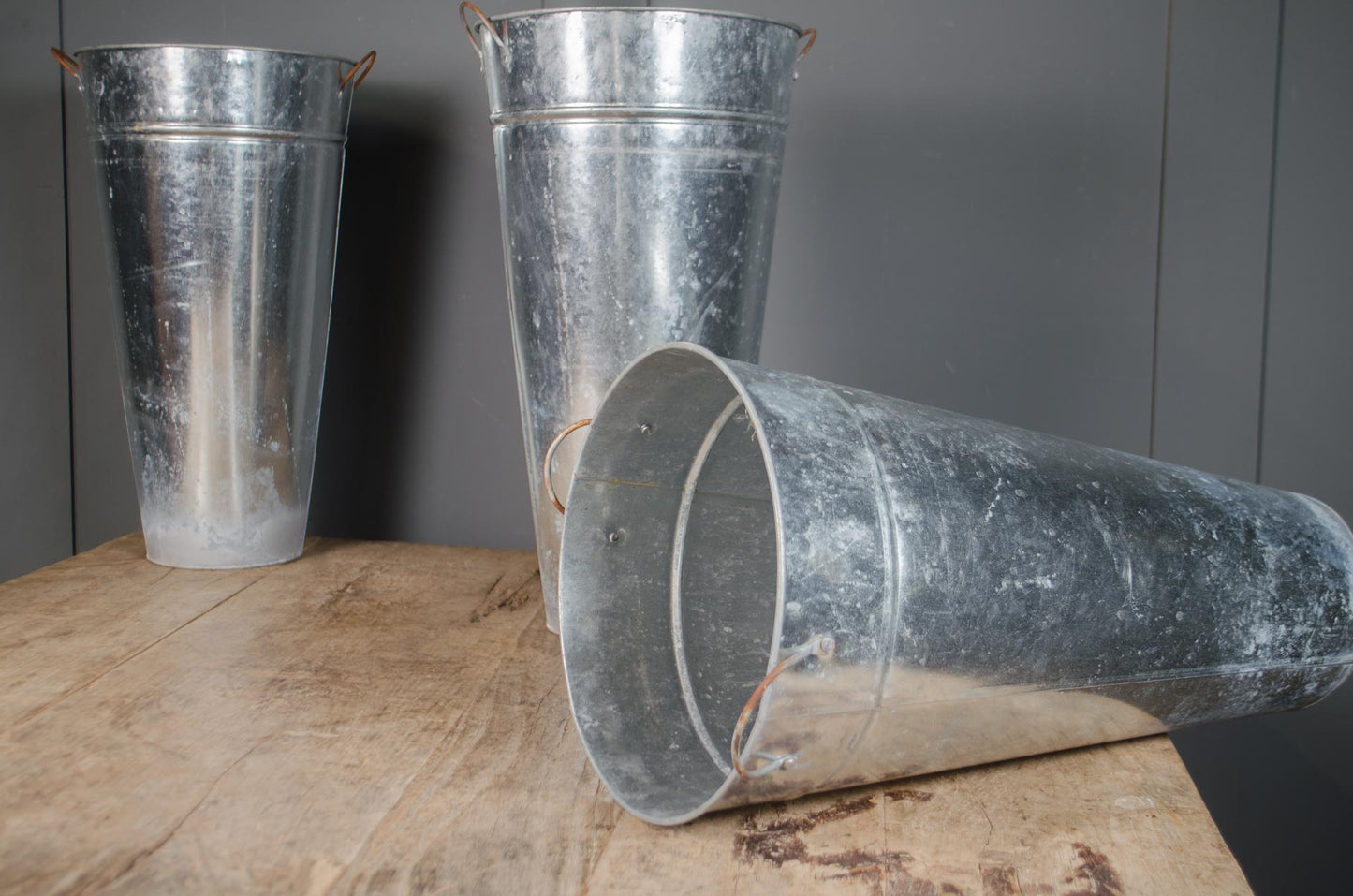 Three galvanized metal buckets in different positions on a rustic wooden table against a dark gray paneled wall.