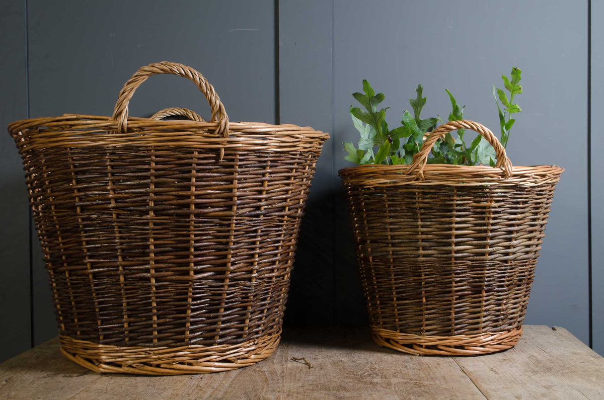 Two natural woven wicker baskets with handles, one large and one small, displayed on a rustic wooden surface against a dark gray paneled wall. The smaller basket is shown holding green leafy stems.