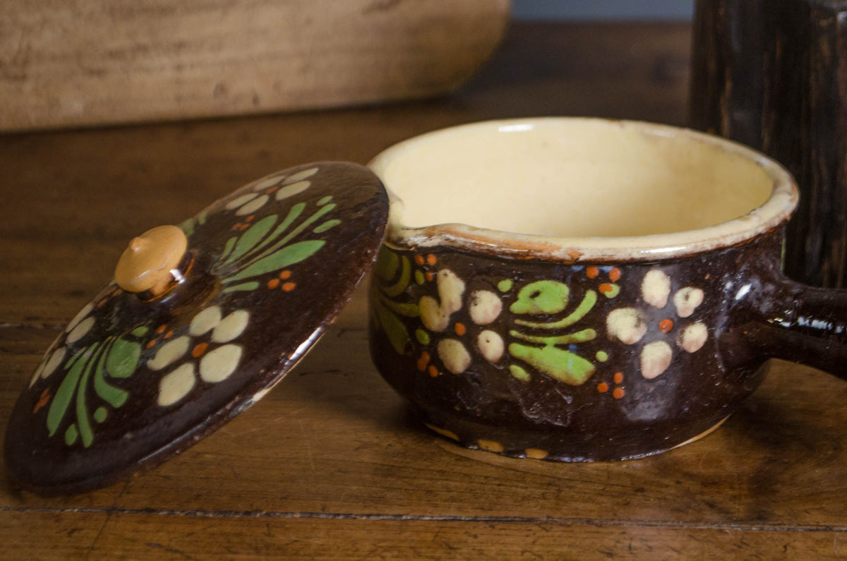 A dark brown ceramic lidded pot with a cream-colored interior and hand-painted green, white, and orange floral motifs, shown on a rustic wooden surface with its lid leaning beside it.
