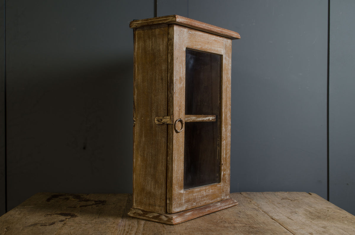 A rustic wooden display cabinet with a glass door and metal latch, standing on a wooden table against a dark gray paneled wall.