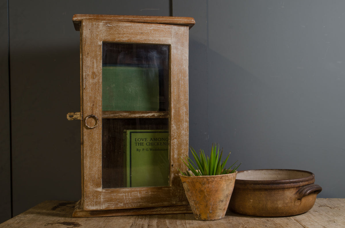 A rustic, weathered light brown wooden cabinet with a glass door and a small brass handle, styled on a wooden table next to a small potted succulent and a ceramic bowl against a dark gray background.