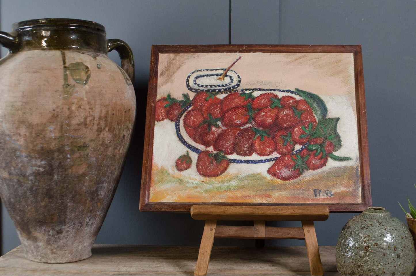 A framed still life painting of red strawberries on a table, displayed on a small wooden easel. The painting is flanked by a large rustic brown ceramic vase on the left and a small speckled pot on the right, set against a dark gray wall.