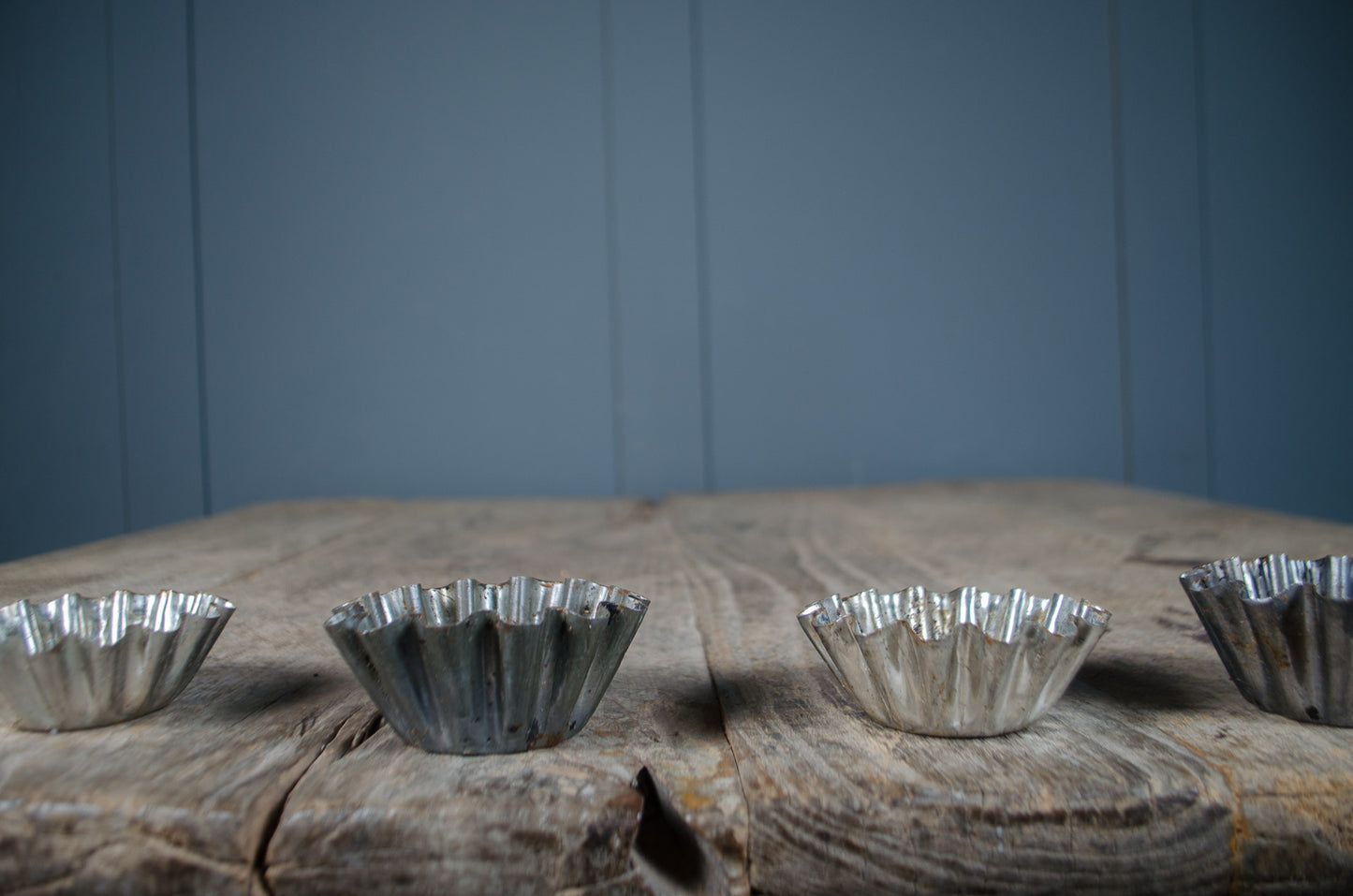Four small silver metal fluted baking cups lined up on a rustic wooden surface against a blue paneled background.