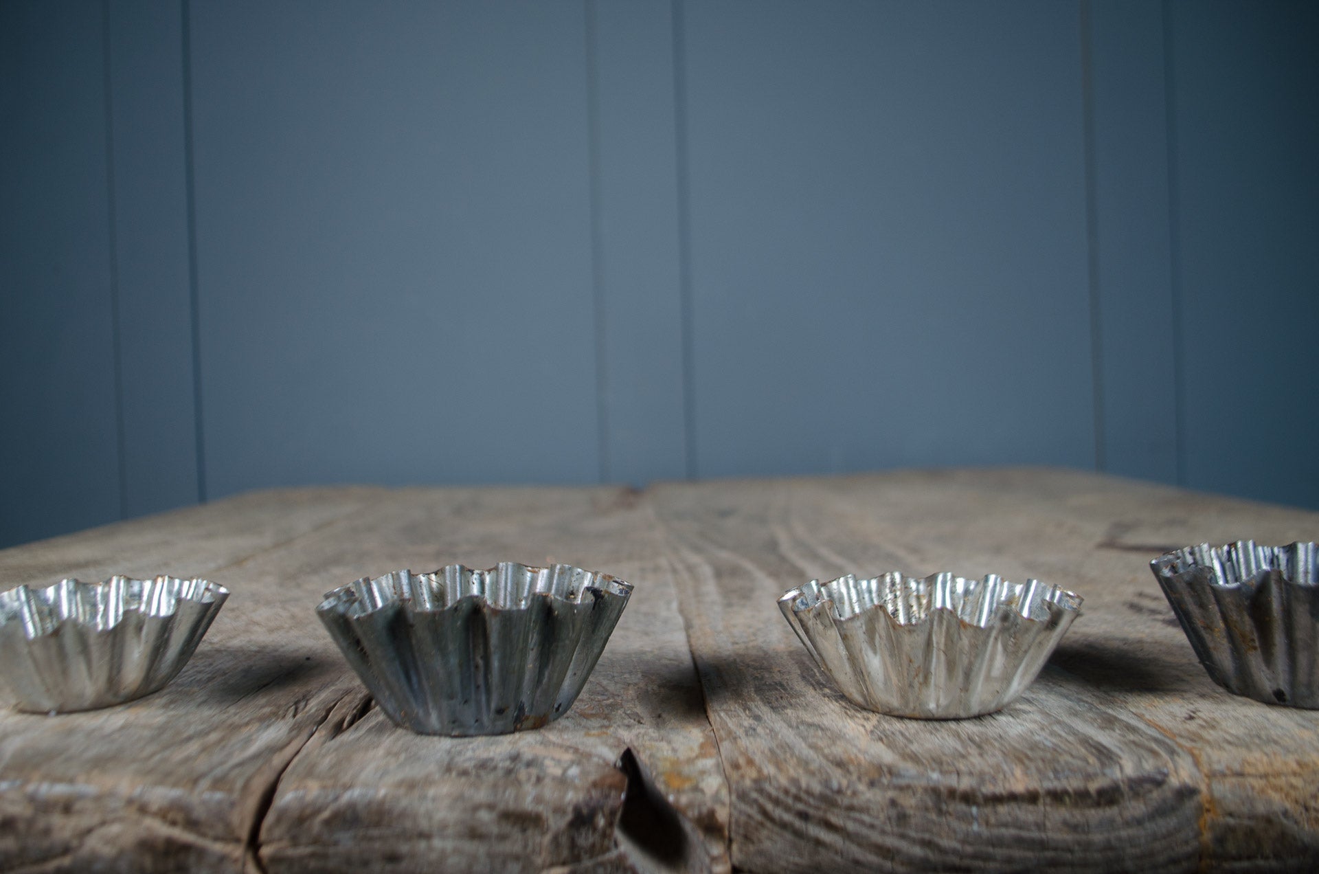 
Four small silver metal fluted baking cups lined up on a rustic wooden surface against a blue paneled background.