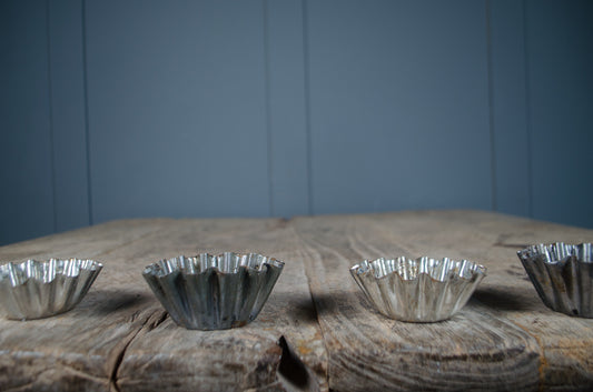 Four small silver metal fluted baking cups lined up on a rustic wooden surface against a blue paneled background.