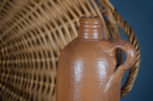 A close-up of a rustic brown clay jug with a handle, positioned in front of a woven wicker basket against a dark blue background.
