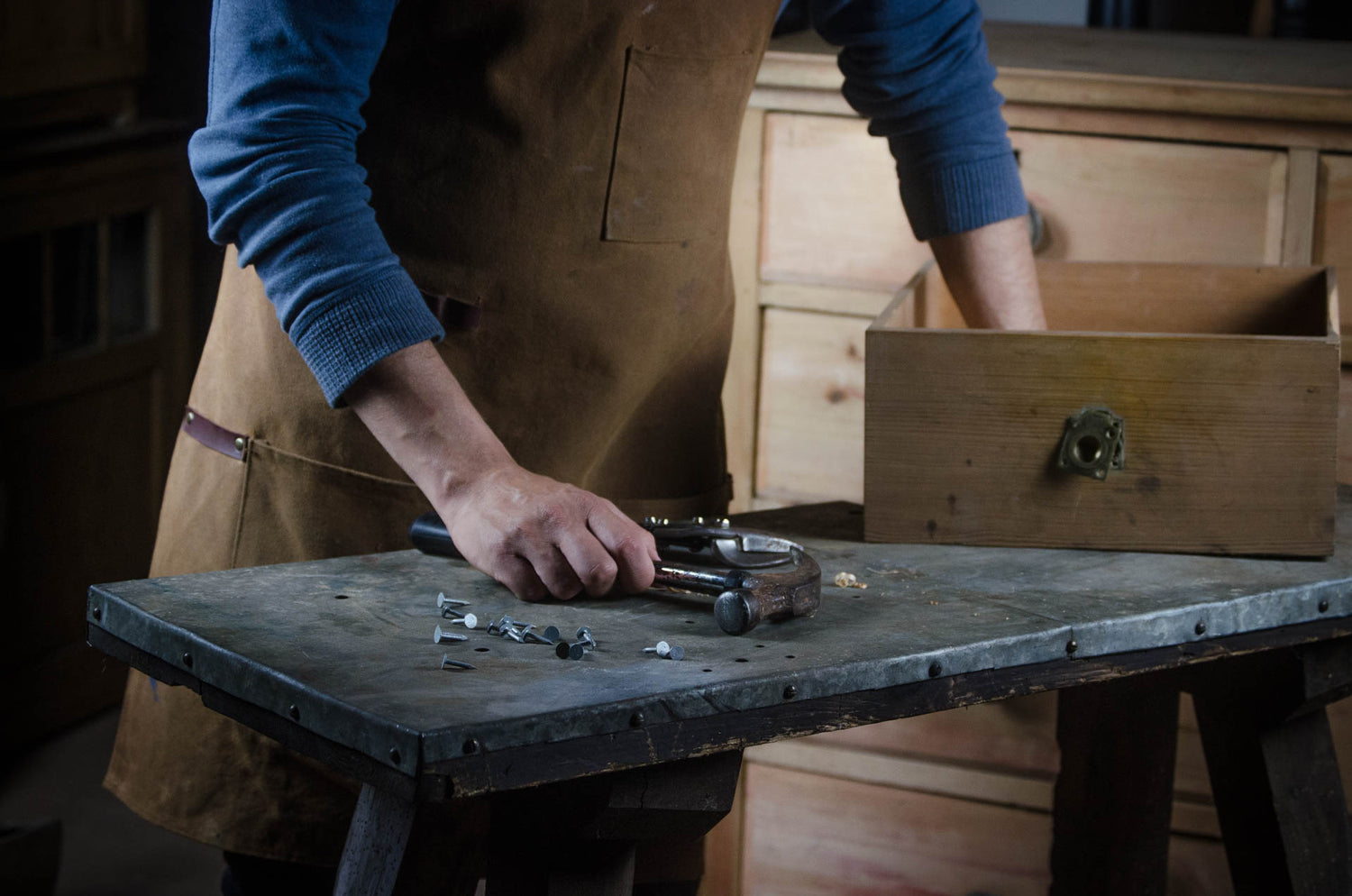 A person wearing a brown leather apron and blue long-sleeve shirt works at a wooden workbench, using a tool on small metal pieces. An open wooden drawer sits on the bench in a workshop setting. Relish lifestyle Devon