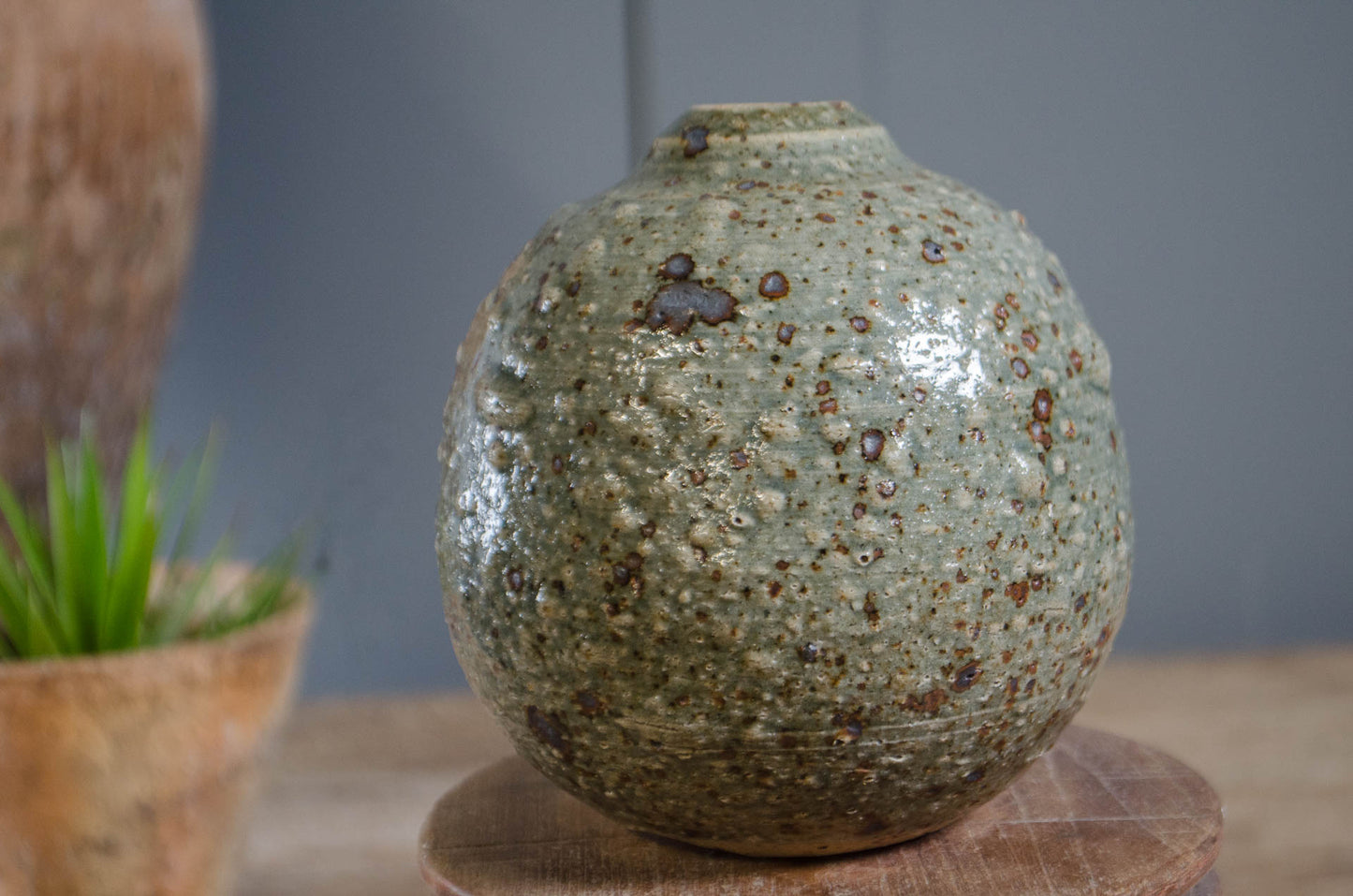 A handmade ceramic vase with a rounded, textured body and a short neck, featuring a speckled glaze in olive green, gray, and brown. It is displayed on a round wooden pedestal with a blurred background containing a small potted plant.
