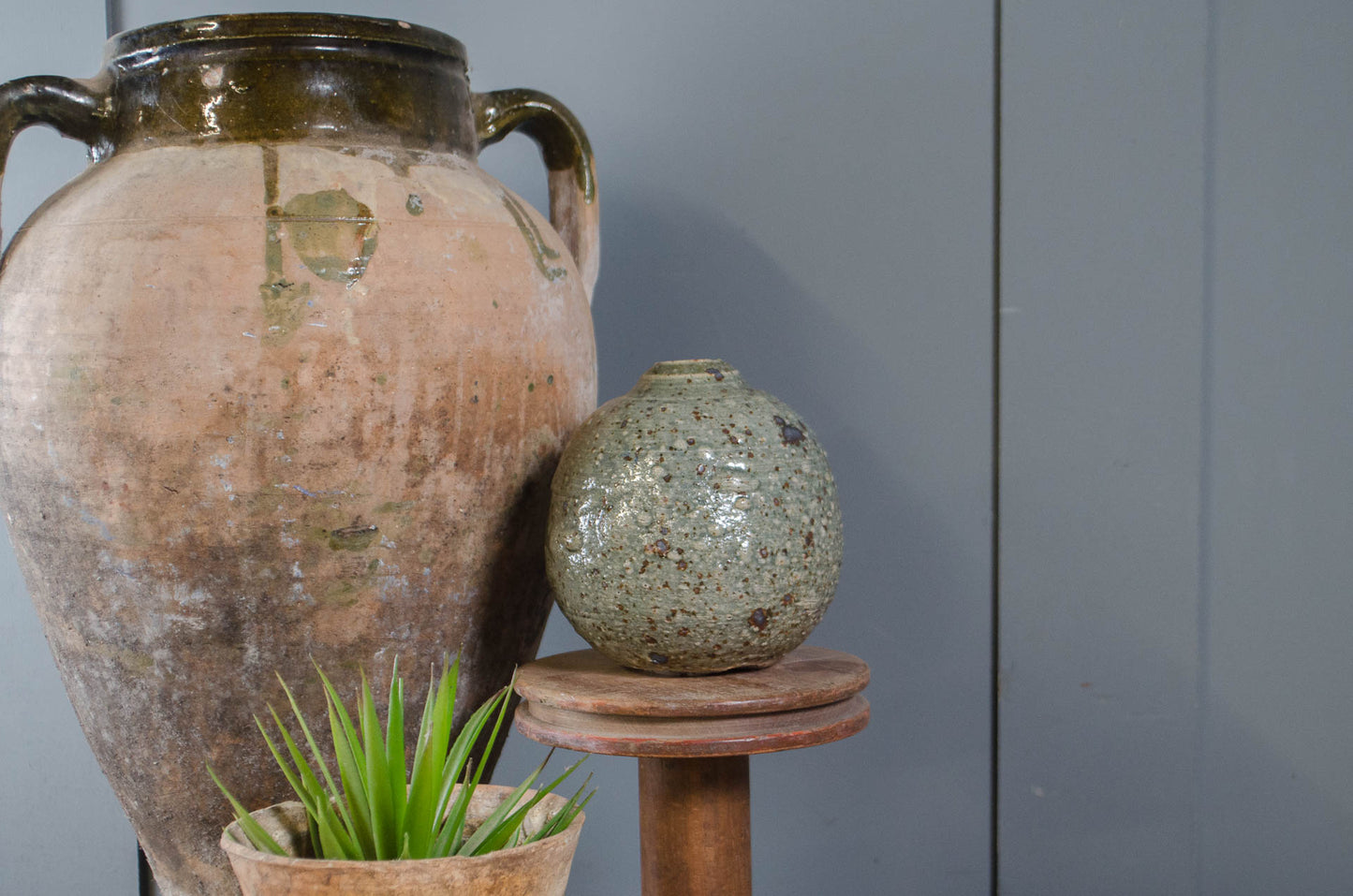 A rustic handmade ceramic set featuring a large brown and green glazed amphora-style vase, a smaller speckled green-gray globular vase on a wooden stand, and a small potted succulent against a gray paneled wall.