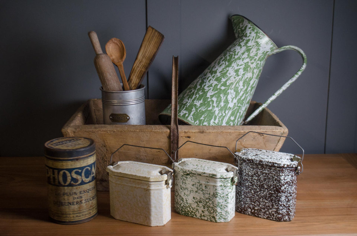 A styled collection of vintage enamel storage containers and kitchenware, including a green and white speckled enamel pitcher, a green speckled enamel canister, a cream speckled enamel canister, a dark brown speckled enamel canister, and a blue vintage canister, arranged on a wooden table against a dark gray paneled wall.