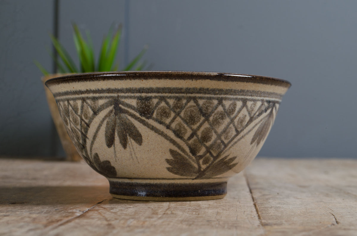 
A handmade ceramic bowl with a beige and brown floral pattern, sitting on a rustic wooden surface with a blurred green plant in the background.