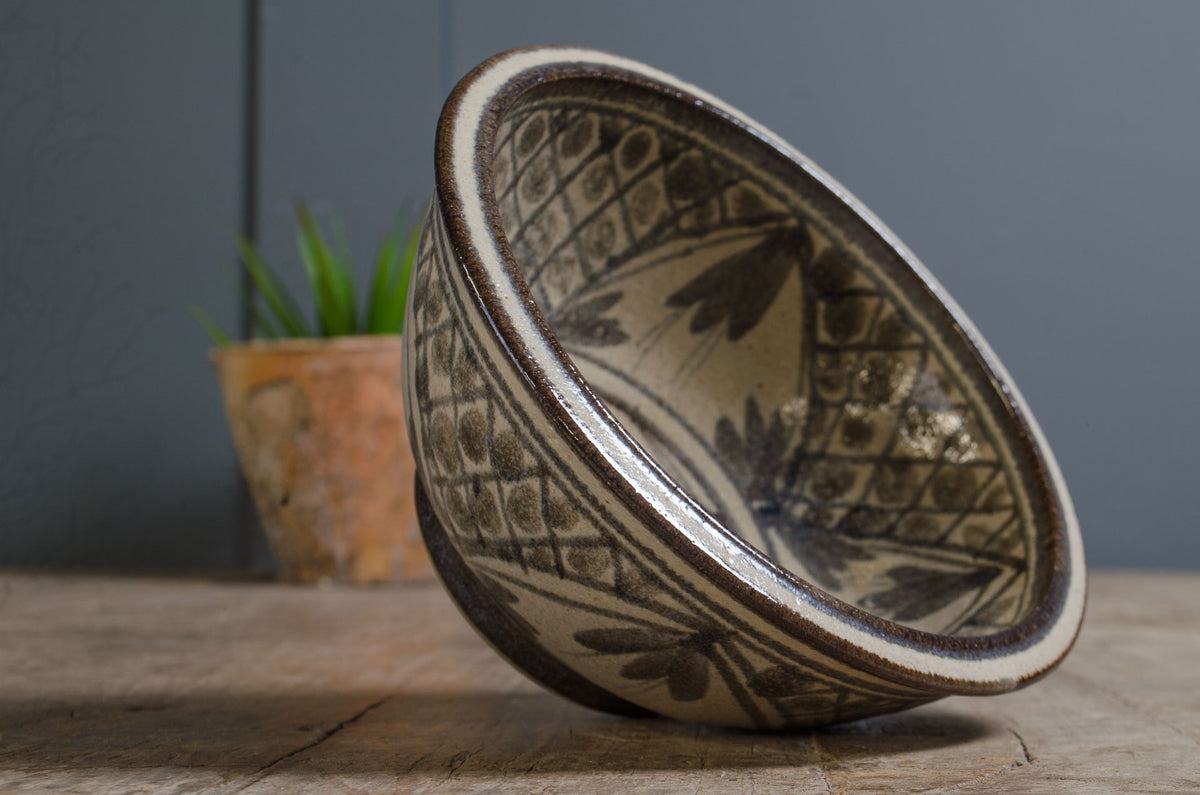 
A handmade ceramic bowl with a rustic hand-painted pattern, resting on a wooden surface against a dark gray background with a blurred potted plant.