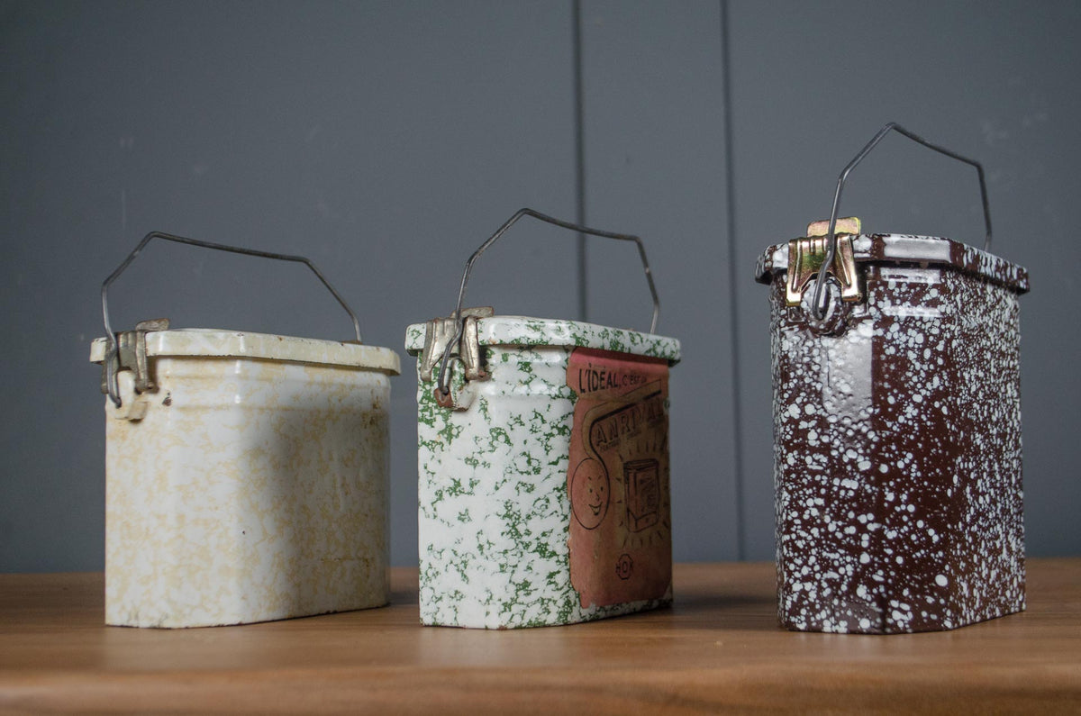 
Three vintage enamel storage boxes with metal handles sit on a wooden surface against a dark gray paneled wall. The left box is cream and beige speckled, the middle is white and green speckled with a brown paper label, and the right box is dark brown and white speckled.
