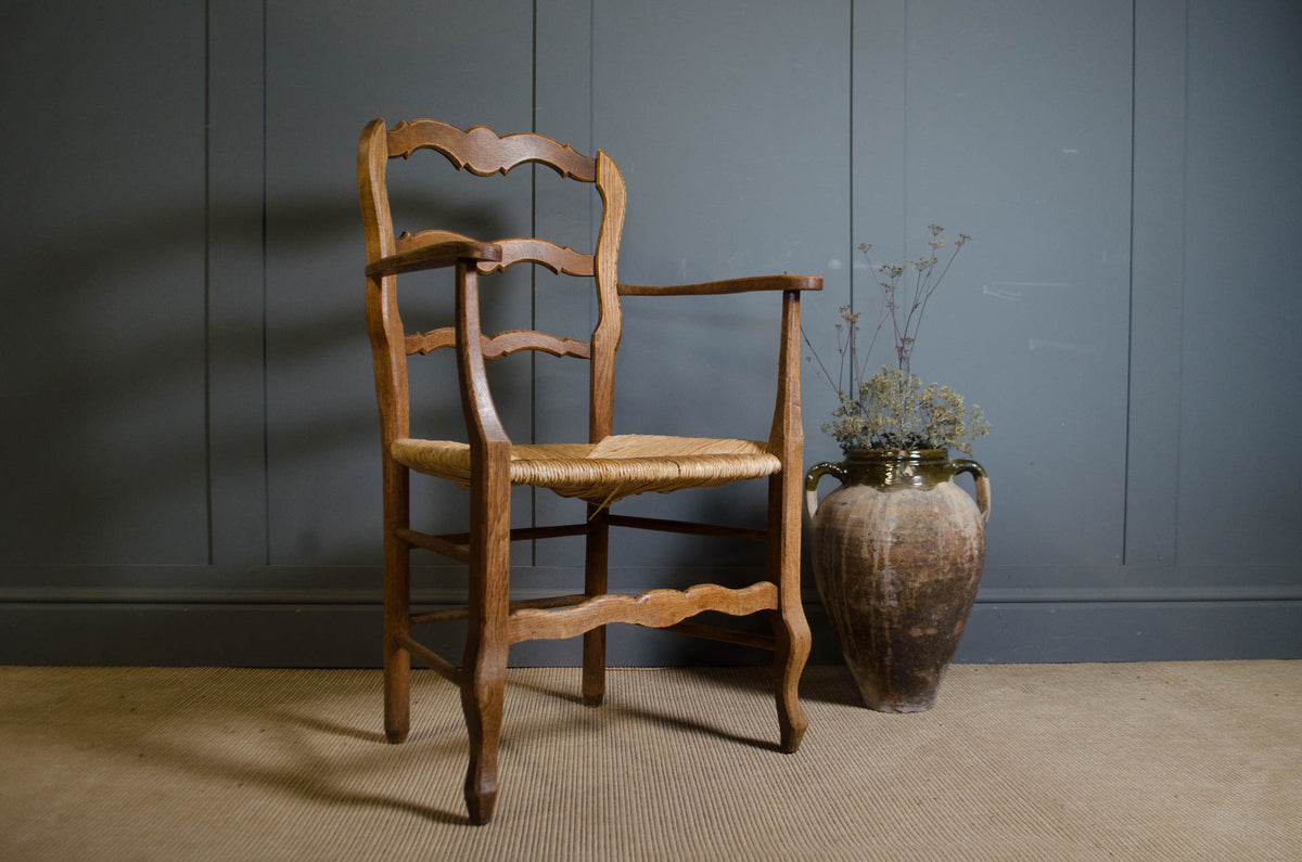 
A rustic wooden ladder-back armchair with a woven seat, positioned next to a large brown ceramic vase with dried branches against a dark gray paneled wall.